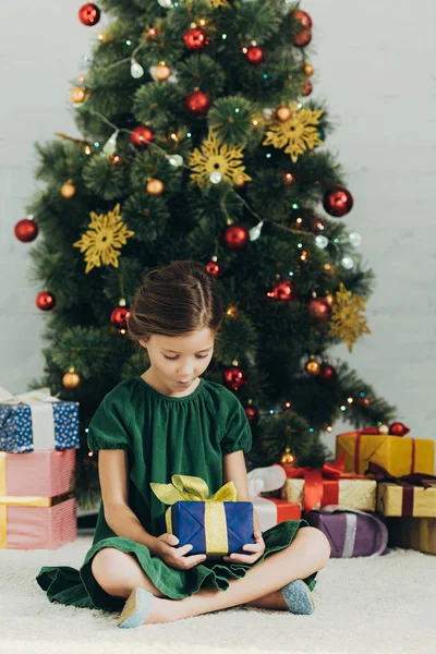 Adorable kid sitting on floor near christmas tree and holding gift box — Stock Photo