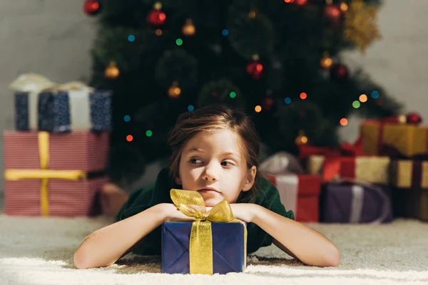 Dreamy child looking away while lying on floor near gift box and christmas tree — Stock Photo