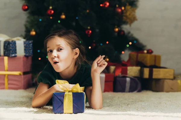 Amüsiertes Kind zeigt mit dem Finger auf Geschenkboxen, während es auf dem Boden in der Nähe des Weihnachtsbaums liegt — Stockfoto