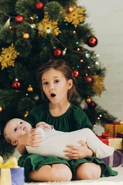 Amused child looking at camera while sitting near christmas tree and holding cute little sister — Stock Photo