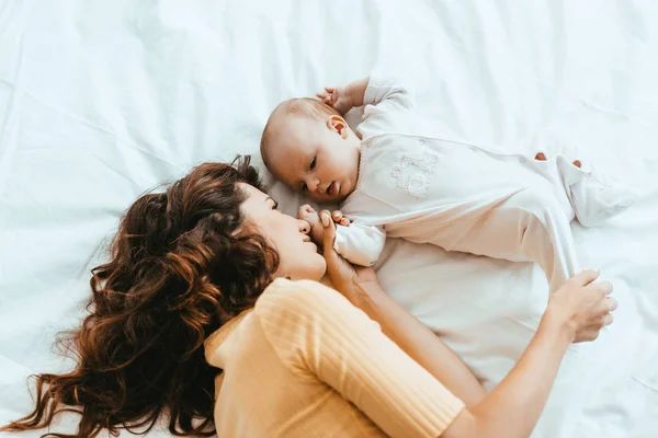 Top view of mother touching leg of cute little daughter while lying on white bedding — Stock Photo