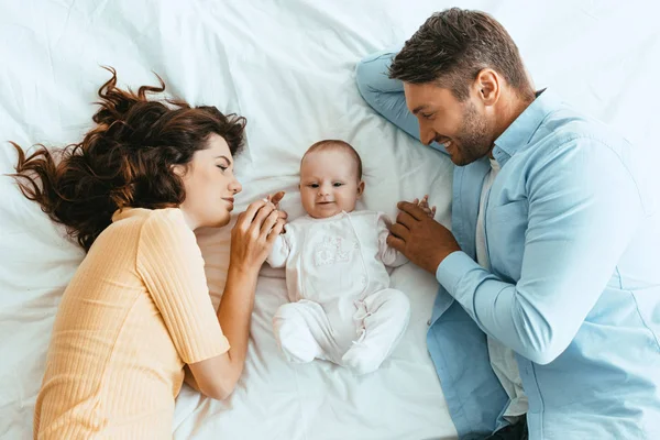 Feliz mamá y papá suavemente tocando adorable bebé mientras están acostados en ropa de cama blanca juntos - foto de stock