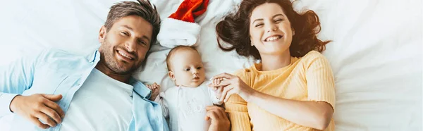 Panoramic shot of happy parents lying on bedding near cute little daughter — Stock Photo