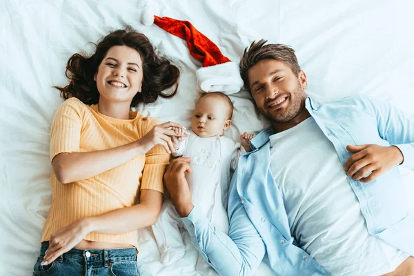 Top view of happy parents lying on bedding near baby and santa hat — Stock Photo