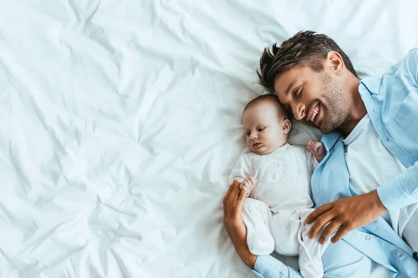 Ansicht von oben: glücklicher Vater umarmt entzückendes Baby, während es auf weißem Bettzeug liegt — Stockfoto