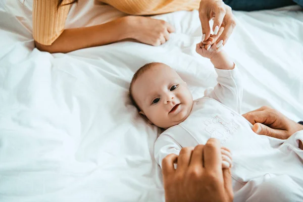 Cropped view of mother and dad playing with smiling baby lying on white bedding — Stock Photo