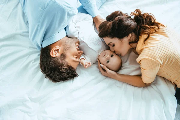 Счастливая мать целует ребенка, лежа на кровати рядом с улыбающимся мужем — стоковое фото