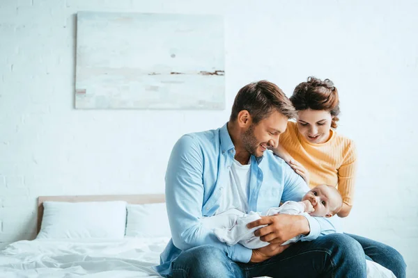 Feliz hombre sosteniendo pequeño bebé mientras está sentado en la cama cerca de sonriente esposa - foto de stock