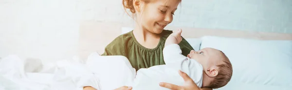 Panoramic shot of happy child holding cute little sister while sitting on bed — Stock Photo