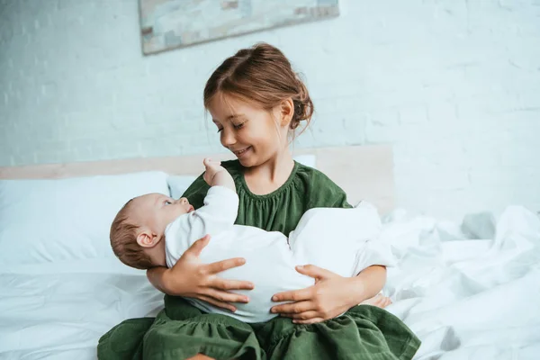 Niño feliz sosteniendo linda hermana pequeña mientras está sentado en la ropa de cama blanca - foto de stock