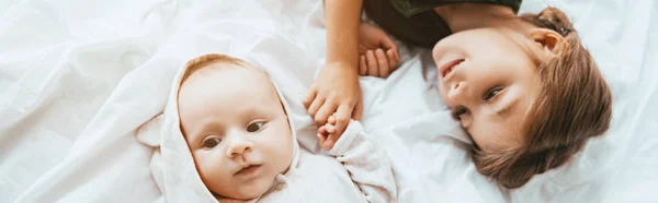 Colpo panoramico di bambino sorridente che tiene la mano della sorellina sdraiata su biancheria da letto bianca — Foto stock