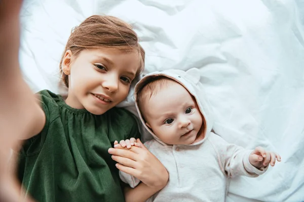 Vista superior del niño sonriente mirando a la cámara mientras está acostado en la cama blanca cerca del bebé lindo - foto de stock