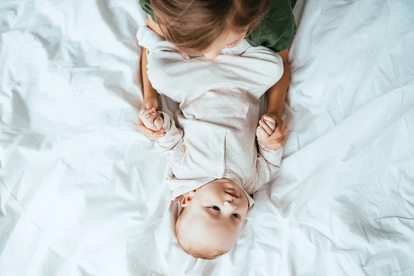 Top view of child holding hands of adorable baby lying on white bedding — Stock Photo