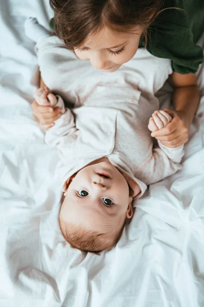 Draufsicht des Kindes, das die Hände eines entzückenden Babys hält, das auf weißem Bettzeug liegt — Stockfoto