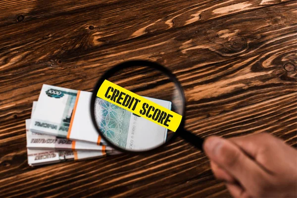 Cropped view of man holding magnifier near russian money on wooden table with credit score illustration — Stock Photo