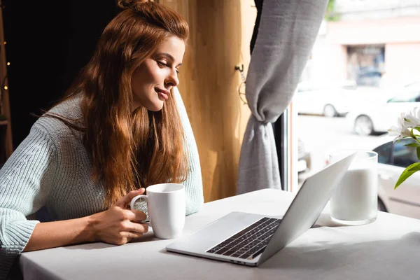 Redhead woman with cup of coffee using laptop in cafe — Stock Photo