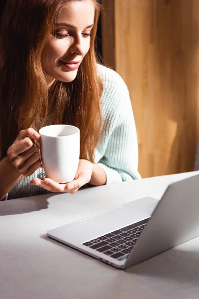 Улыбающаяся рыжая женщина с чашкой кофе с помощью ноутбука в кафе — стоковое фото