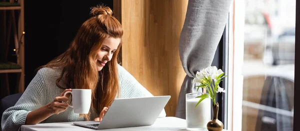 Hermosa mujer sonriente con taza de café usando el ordenador portátil en la cafetería - foto de stock