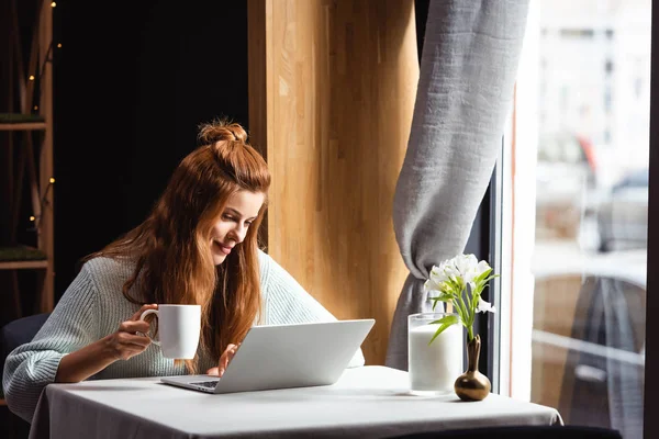 Mujer pelirroja sonriente con taza de café usando el ordenador portátil en la cafetería - foto de stock