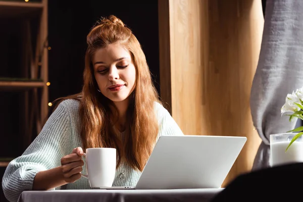 Pelirroja chica pensativa con taza de café utilizando el ordenador portátil en la cafetería - foto de stock