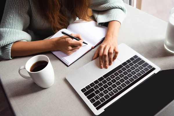 Vista cortada da mulher escrevendo no bloco de notas e usando laptop no café com xícara de café — Fotografia de Stock