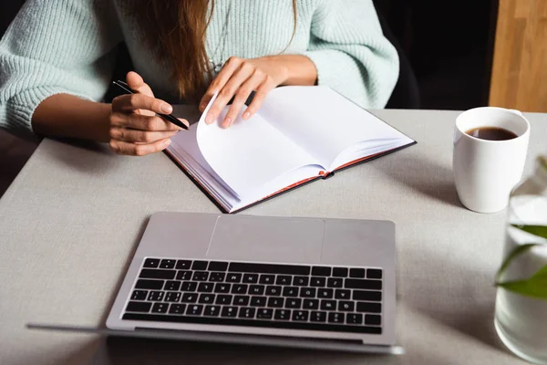 Обрезанный вид женщины, обучающейся онлайн с блокнотом и ноутбуком в кафе с чашкой кофе — стоковое фото