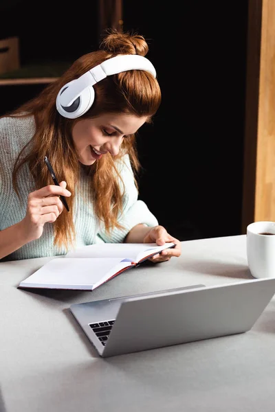 Mulher feliz em fones de ouvido escrever e estudar on-line com laptop no café — Fotografia de Stock