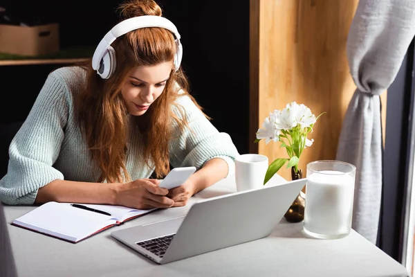 Концентрированная женщина в наушниках с помощью смартфона и ноутбука в кафе — стоковое фото