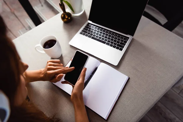 Обрезанный вид женщины, обучающейся онлайн со смартфоном и ноутбуком в кафе с чашкой кофе — стоковое фото