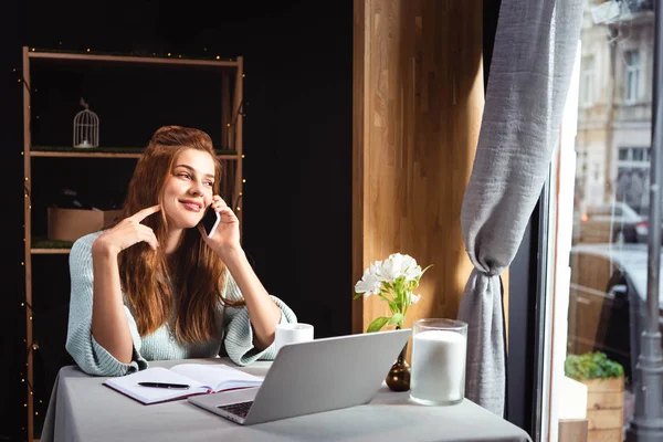Mujer sonriente hablando en el teléfono inteligente mientras trabaja en el ordenador portátil en la cafetería - foto de stock