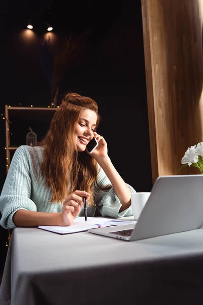 Mujer sonriente hablando en el teléfono inteligente mientras estudia en línea con el ordenador portátil en la cafetería - foto de stock