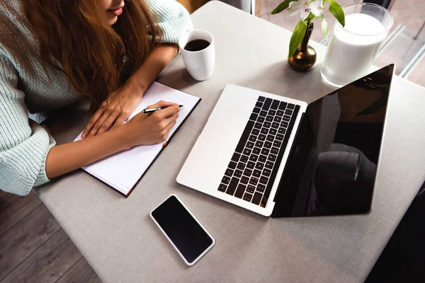 Vista cortada da mulher escrevendo no bloco de notas no café com smartphone e laptop — Fotografia de Stock