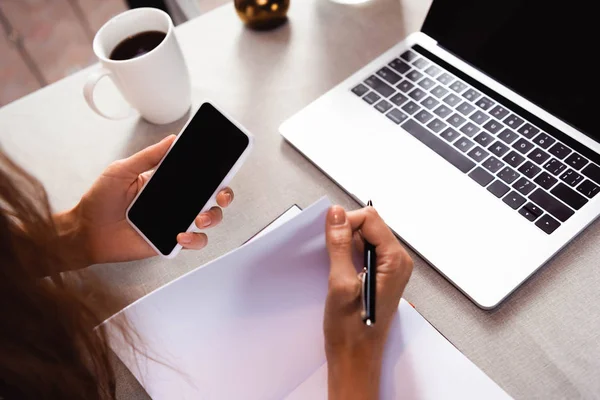 Vista ritagliata di ragazza che lavora con blocco note, smartphone e laptop in caffè — Foto stock