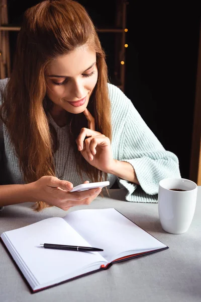 Mulher atraente usando smartphone no café com bloco de notas e café — Fotografia de Stock