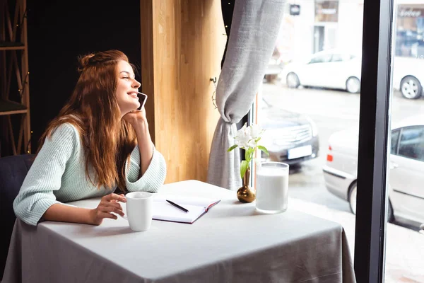 Mulher feliz atraente falando no smartphone no café com bloco de notas e xícara de café — Fotografia de Stock