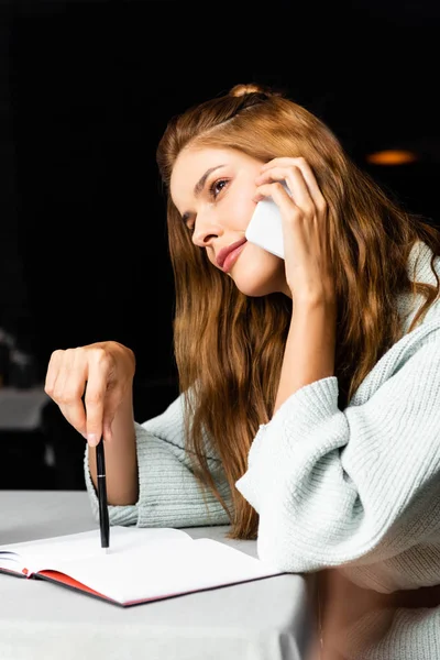 Привлекательная задумчивая женщина разговаривает на смартфоне в кафе с блокнотом — стоковое фото