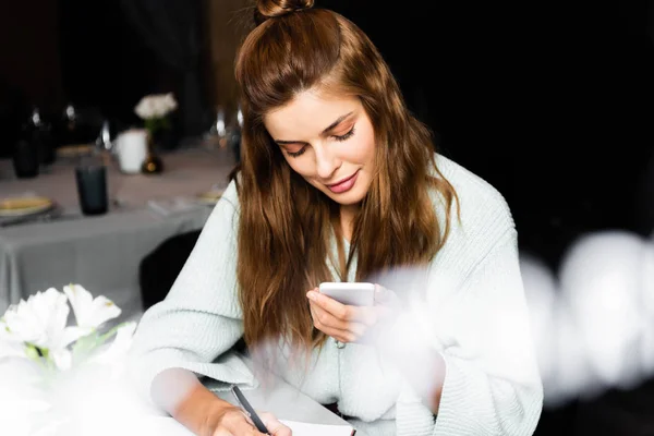 Belle femme utilisant smartphone et écriture dans le bloc-notes dans le café avec une tasse de café — Photo de stock