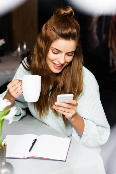 Happy woman with cup of coffee using smartphone in cafe with notepad — Stock Photo