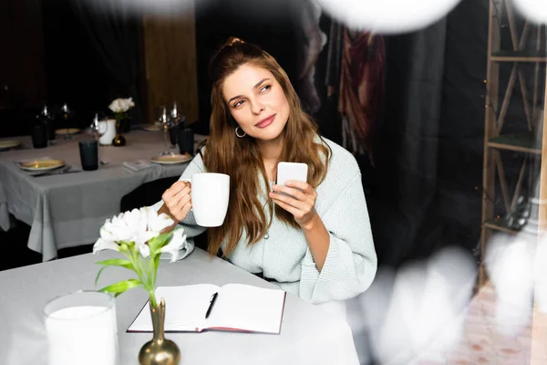 Мечтательная женщина с чашкой кофе с помощью смартфона в кафе с блокнотом — стоковое фото
