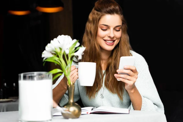 Smiling woman with cup of coffee using smartphone in cafe with notepad — Stock Photo