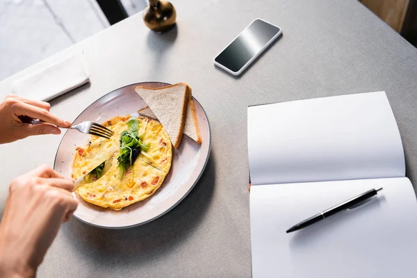 Cropped view of woman eating omelet in cafe with empty notepad and smartphone — Stock Photo
