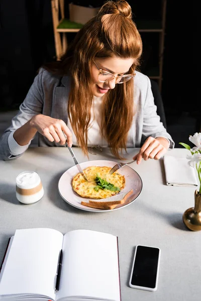 Pelirroja comiendo tortilla para el desayuno con café en la cafetería con bloc de notas vacío y teléfono inteligente - foto de stock