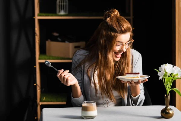 Mujer emocionada comiendo pastel con café en la cafetería con flores en jarrón - foto de stock