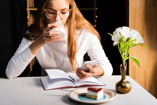 Привлекательная девушка пьет кофе при использовании смартфона в кафе с блокнотом, тортом и цветами — стоковое фото