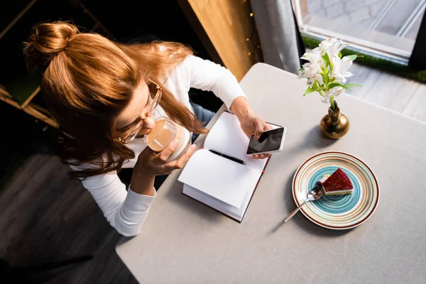 Вид зверху на руду жінку, яка п'є каву, використовуючи смартфон у кафе з блокнотом, тортами та квітами — стокове фото