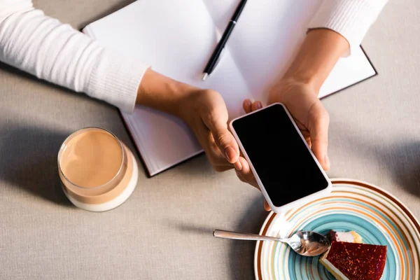 Cropped view of woman with notepad using smartphone in cafe with cake and coffee — Stock Photo