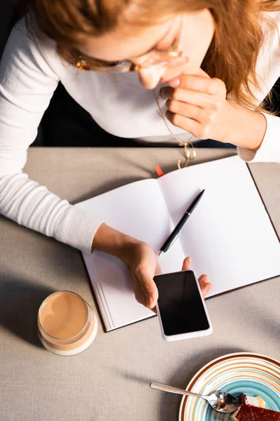 Overhead view of woman with notepad using smartphone in cafe with glass of coffee — Stock Photo