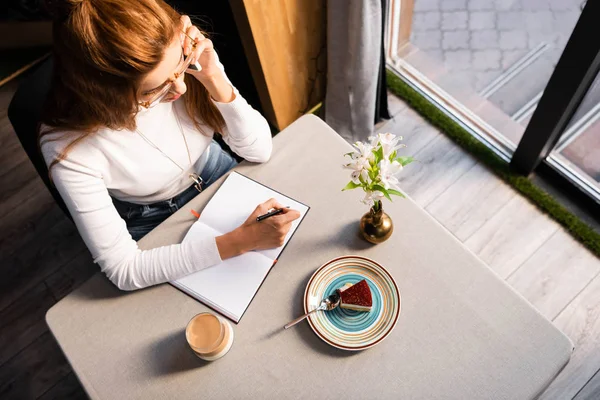 Над головой вид женщины, пишущей в блокноте и разговаривающей на смартфоне в кафе с тортом и кофе — стоковое фото