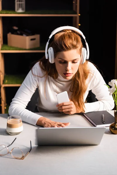 Beautiful focused woman in headphones watching webinar on laptop in cafe with smartphone — Stock Photo
