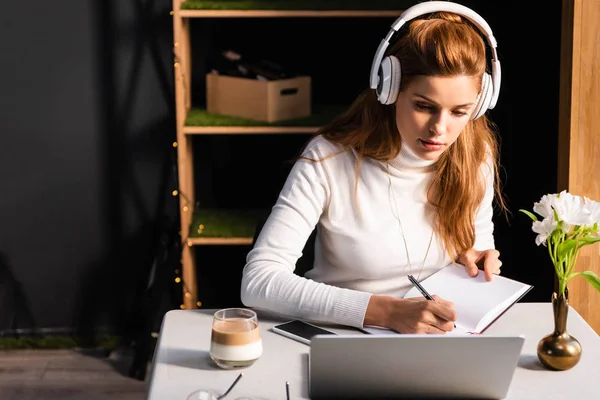 Beautiful redhead woman in headphones writing in notepad while watching webinar on laptop in cafe — Stock Photo